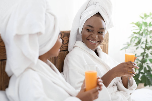 Two joyful black women drinking fresh juice in bed - Фото, изображение