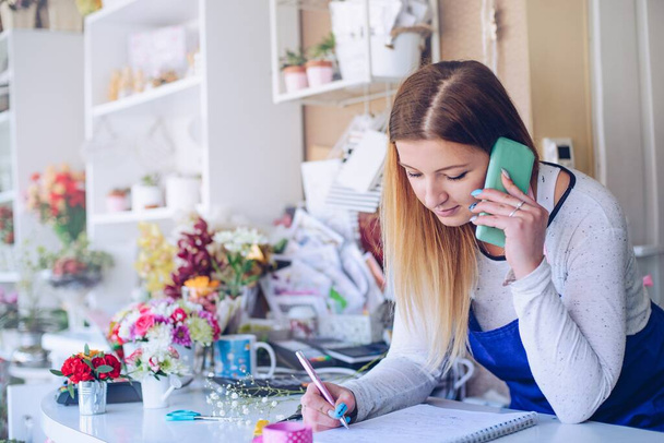 Молода жінка-флорист відзначає замовлення клієнта під час розмови мобільних телефонів у квітковому магазині
 - Фото, зображення