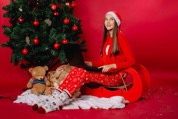 Une fille dans un chapeau du Père Noël sur un traîneau, à la maison près d'un arbre de Noël avec un ordinateur portable. Photo de haute qualité - Photo, image