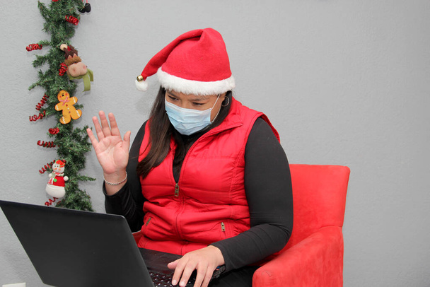 mujer latina con máscara de protección, sombrero de santa claus y portátil en videoconferencia, decoración de Navidad, nuevo covid-19 normal - Foto, Imagen