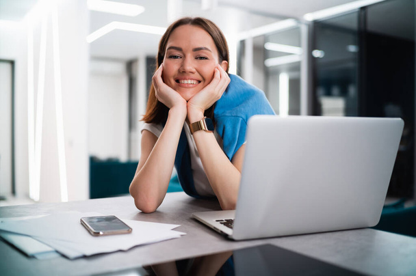 Boldog mosolygós fiatal nő ül az asztalnál nyitott laptoppal a kamerába nézve. Sikeres mosolygós diáklány otthon marad az online tanulási eszközök és platformok segítségével. - Fotó, kép
