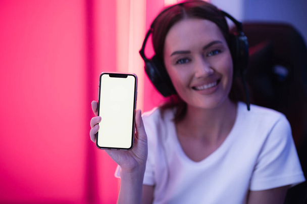 Bonne gamer fille portant casque montrant l'affichage du téléphone intelligent et pointant vers l'écran vide avec son doigt. - Photo, image