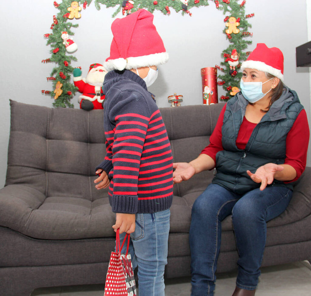 Grootmoeder en kind met beschermkapje en kerstmuts, cadeaus en kerstversiering, nieuwe normale covid-19 - Foto, afbeelding