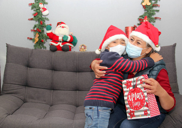 grandmother and child with protection mask and santa claus hat, gifts and Christmas decorations, new normal covid-19 - Photo, Image
