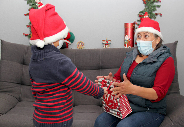 babička a dítě s ochrannou maskou a Santa Claus kloboukem, dárky a vánoční výzdobou, nový normální covid-19 - Fotografie, Obrázek