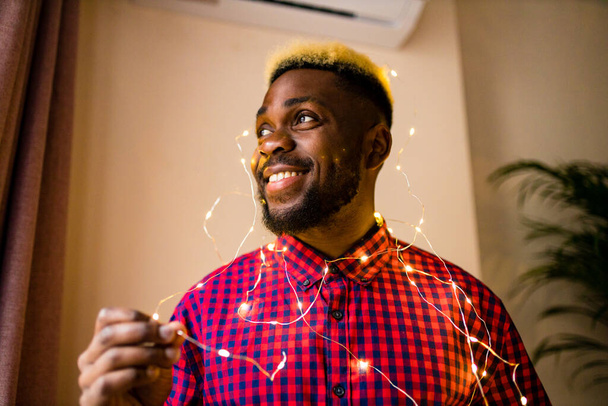 african man making preparations for Christmas party and decorating home with glowing garland - Foto, imagen