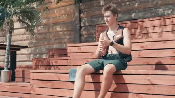 夏の日に太陽が凍る間、スポーツボトルから水をすすりながら木製のベンチに座っているウォータースポーツライフベストの若い男性サーファーの完全な映像 - 映像、動画