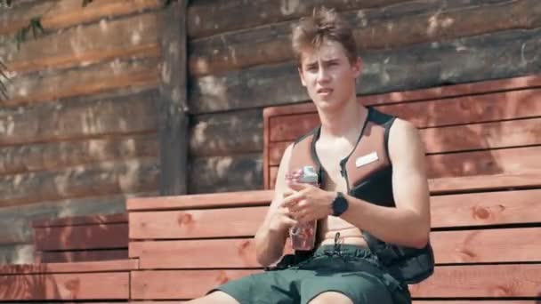 Mittlere Aufnahme eines kaukasischen Jungen in Wassersport-Schwimmweste, der Wasser trinkt, während er sich an einem heißen Sommertag auf einer Bank entspannt - Filmmaterial, Video