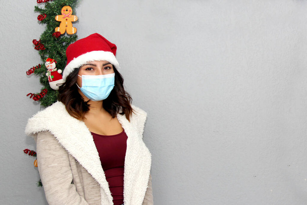 Латинська жінка з маскою захисту, светром і різдвяним капелюхом на сірому фоні з різдвяною прикрасою, нова нормальна
 - Фото, зображення