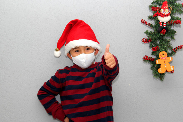 7χρονο λατίνο αγόρι με μάσκα προστασίας, πουλόβερ και χριστουγεννιάτικο καπέλο σε γκρι φόντο με χριστουγεννιάτικη διακόσμηση, νέο κανονικό covid-19 - Φωτογραφία, εικόνα