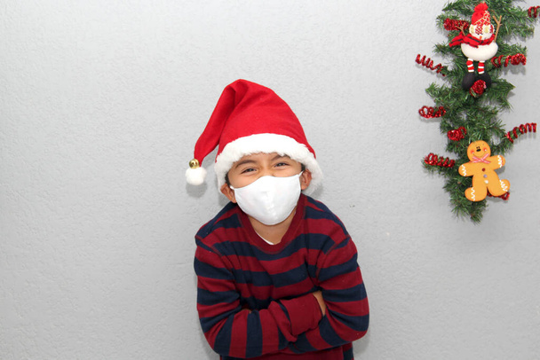 Garçon latin de 7 ans avec masque de protection, pull et chapeau de Noël sur fond gris avec décoration de Noël, nouveau covide normal-19 - Photo, image