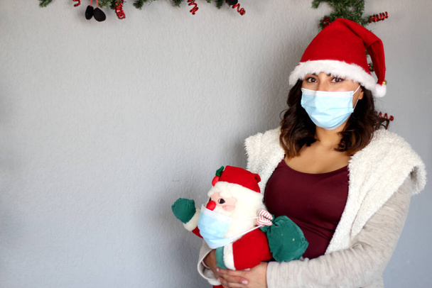保護マスクを持つ若いラテン女性,セーターとサンタクラスの帽子,クリスマスの装飾とサンタクラスの人形,新しい通常のcovid-19 - 写真・画像