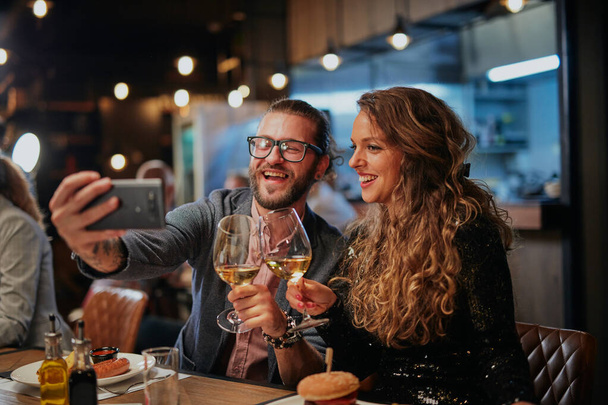 Atractiva pareja hipster sentada en un restaurante y tomando una selfie. Es hora de cenar.. - Foto, Imagen