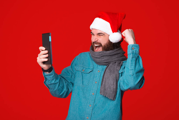 クリスマスの帽子をかぶって興奮した髭の勝者は彼のタブレットを見ています。 - 写真・画像