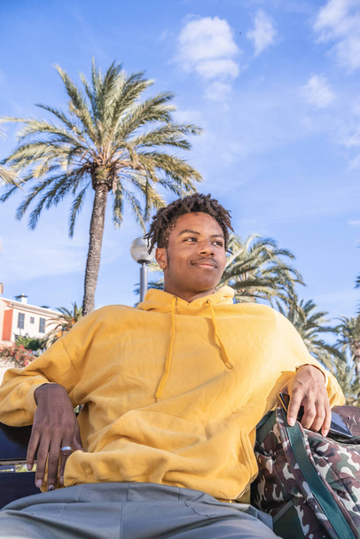 afro american black man with yellow sweatshirt looking at smartphone laying on a wooden bench in a park - Фото, зображення
