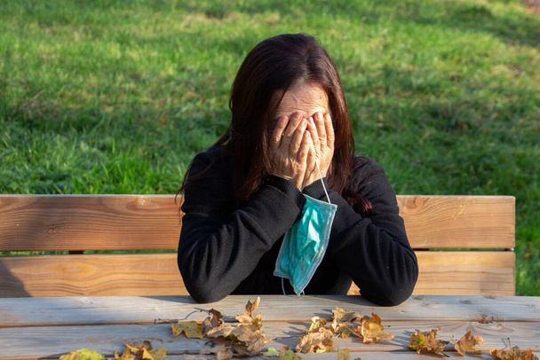 Woman with dark hair sitting at a picnic table holding a medical mask and her head down on her hands. - Photo, Image