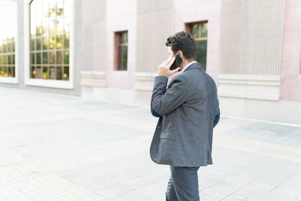 Вид сзади на профессионала в блейзере, идущего и разговаривающего по телефону. Взрослый мужчина с портфолио за пределами городского здания  - Фото, изображение