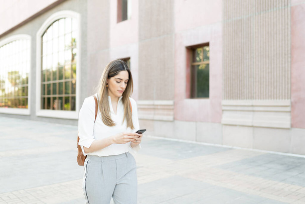 Donna adulta caucasica con uno zaino che cammina e messaggia nel suo smartphone fuori da un edificio urbano  - Foto, immagini