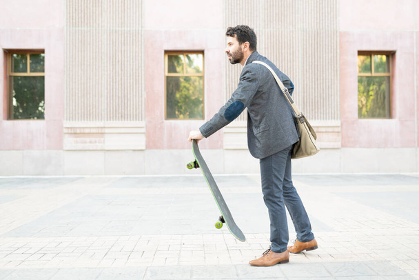 Boční pohled na profesionálního zaměstnance po třicítce, jak se připravuje na jízdu na skateboardu, aby se dostal do práce ve městě  - Fotografie, Obrázek