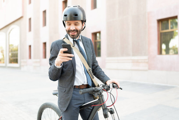 Hombre guapo sonriendo y enviando mensajes de texto en su teléfono inteligente fuera de un edificio de oficinas. Trabajador profesional que utiliza un casco y una bicicleta como vehículo ecológico  - Foto, Imagen