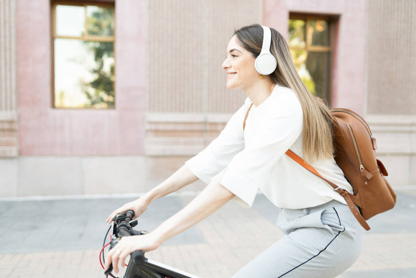 Szczęśliwa i aktywna pracownica ze słuchawkami i plecakiem jeżdżąca na rowerze do pracy  - Zdjęcie, obraz