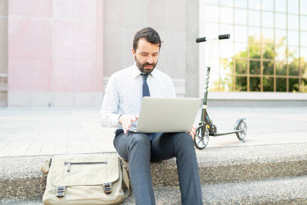 Bussinesman з рюкзаком сидять біля будинку і працюють на своєму ноутбуку поруч з екологічно дружнім скутером - Фото, зображення