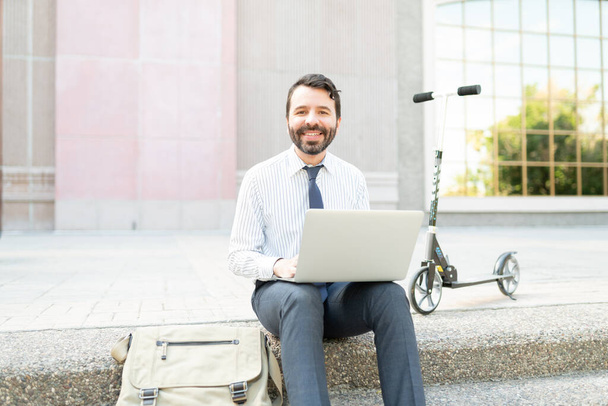 Glücklicher Mann in seinen Dreißigern, der mit einem Roller, einem alternativen Transportmittel, außerhalb des Büros an seinem Computer arbeitet - Foto, Bild