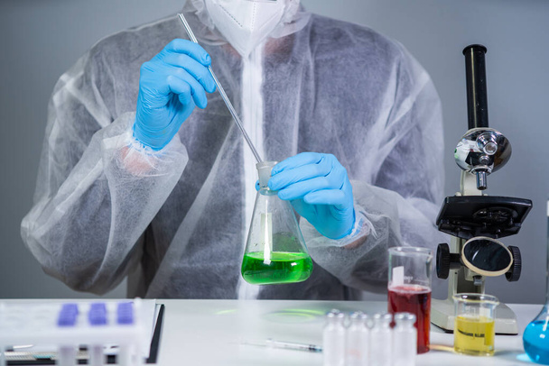 Teknikko suojavaatetuksessa käsi sekoittaen ja ravistaen vihreää nestettä Erlenmeyerin pullossa kemiallisessa laboratoriossa. Laboratorioteknikko tutkii kemiallisia reaktioita ja etsii syöpälääkettä. - Valokuva, kuva