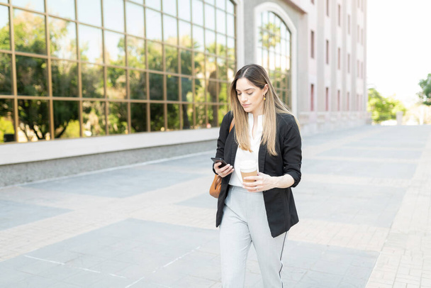 Attraktive kaukasische Frau mit professionellem Outfit, die Kaffee trinkt, zu ihrem Arbeitsplatz läuft und mit dem Smartphone SMS schreibt - Foto, Bild