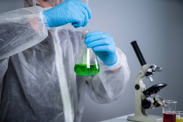 Tutkija ravistelee vihreää biokemiallista tuotetta erlenmeyerpullojen sekoittamisessa ja laboratoriotutkimuksissa. Laboratorioassistentti koronaviruspandemian laboratoriossa, COVID 19. Lääkekehitys, lääketieteellinen tutkimus. - Valokuva, kuva