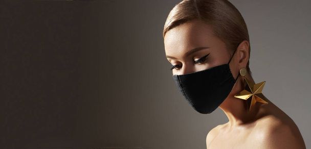 Beautiful woman with a black medicine mask on face. Fashion eye make-up, celebrate style, gold jewelry. Beauty with protection hygiene in viral covid-19 pandemic. Party christmas stylish - Valokuva, kuva