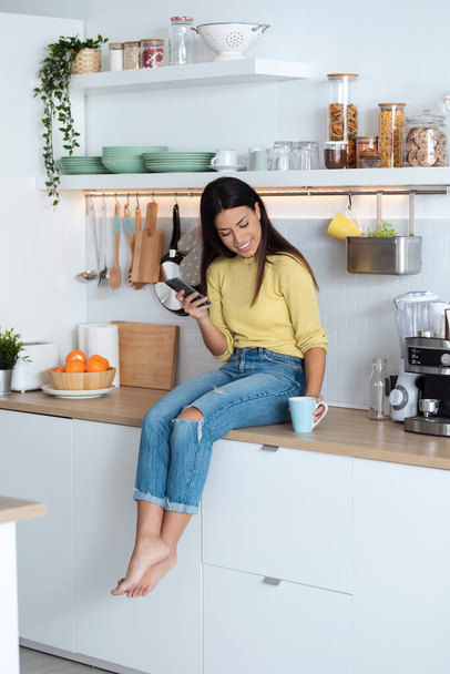 Shot of attractive young woman using smart phone while relaxing drinking coffee sitting on the kitchen at home. - 写真・画像