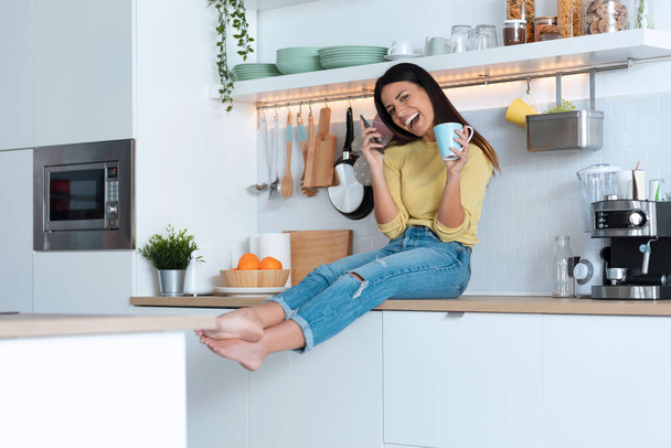 Snímek atraktivní mladá žena dělá vtipné věci, zatímco drží chytrý telefon a šálek kávy sedí na kuchyni doma. - Fotografie, Obrázek
