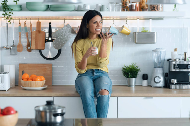 Πυροβολισμός ελκυστική νεαρή γυναίκα τρώει γιαούρτι, ενώ χρησιμοποιεί το έξυπνο τηλέφωνό της κάθεται στην κουζίνα στο σπίτι. - Φωτογραφία, εικόνα