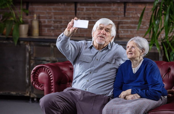 Senior couple taking selfie with smart phone while sitting on couch at home - Foto, afbeelding