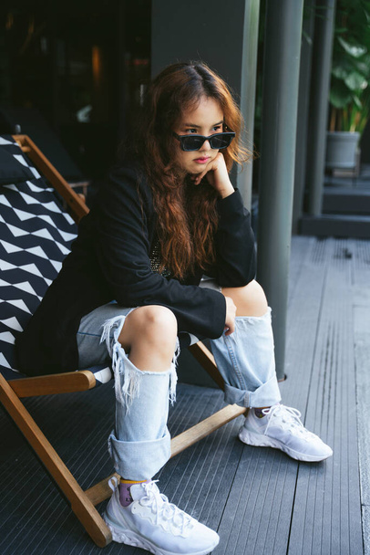 Fashion portrait of young asian woman wearing sunglasses and black jacket sitting on chair beach. - 写真・画像