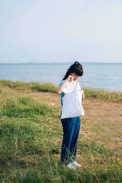 Μαύρο μισό δεμένο κορίτσι μαλλιά σε λευκό και μπλε χρώμα γραβάτα στέκεται σε χορτολιβαδικές εκτάσεις δίπλα στη θάλασσα με τσάντα καμβά. - Φωτογραφία, εικόνα