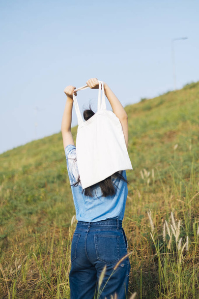Back shot of black half tied hair girl in white and blue tie dye standing in slope grassland and raise the canvas bag up. - Photo, Image