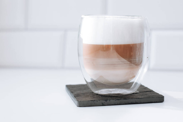 Cappuccino coffee in a double glass on a black slate board against white bricks wall background. - Foto, Imagen