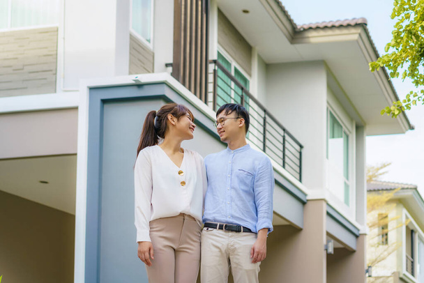 Portrait of Asian young couple standing and hugging together looking happy in front of their new house to start new life. Family, age, home, real estate and people concept. - 写真・画像
