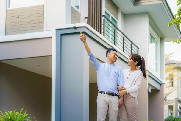 Portrait of Asian couple walking hugging and pointing together looking happy in front of their new house to start new life. Family, age, home, real estate and people concept. - Photo, Image