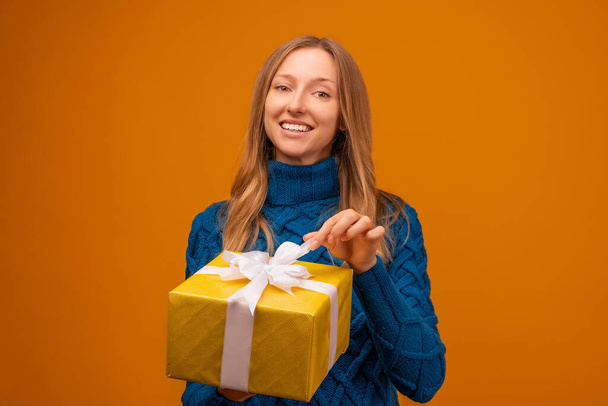 Egy boldog fiatal nő portréja kötött kék pulóverben, kezében ajándékkal, szalaggal díszítve. Stúdiófelvétel, sárga háttér. Újév, Nők napja, Születésnap, Ünnepi koncepció - Fotó, kép