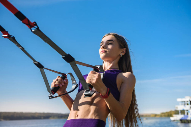 Ładna kobieta w sportowym stroju robi crossfit push upy z trx paskami fitness na świeżym powietrzu w pobliżu jeziora w ciągu dnia. Zdrowy styl życia - Zdjęcie, obraz