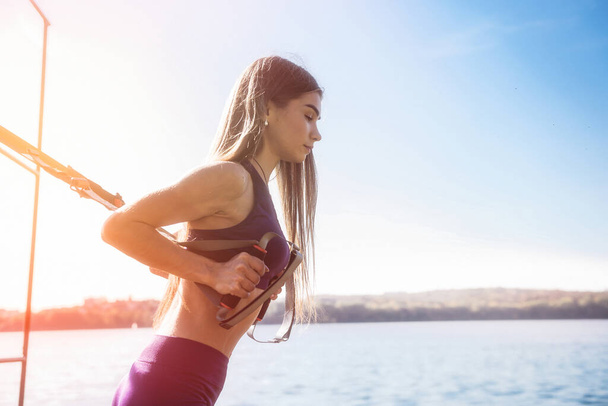 bella donna che esercita con cinghie di sospensione TRX all'aperto vicino al lago durante il giorno. Stile di vita sano - Foto, immagini