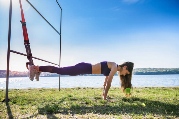 Pretty woman with sportwear doing crossfit push ups with trx fitness straps outdoors near the lake at daytime. Healthy lifestyle - Fotó, kép
