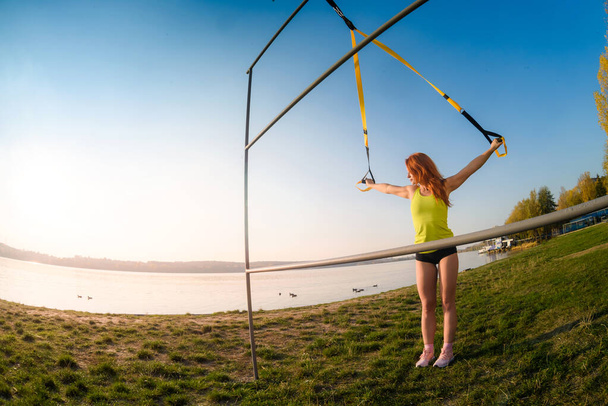 Genç bayan sporcu parkta trx fitness kayışlarıyla antrenman yapıyor. Sağlıklı çalışma ve yaşam tarzı kavramı - Fotoğraf, Görsel