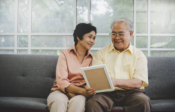 Senior Asian couple is looking at memory photo on a couch at home for happy retirement lifestyle concept - Photo, Image