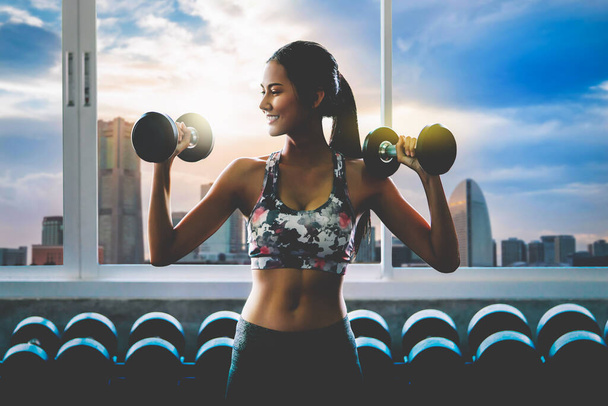 Sportieve vrouw houdt fitness halter met glimlach uit te werken met de moderne stad op de achtergrond - Foto, afbeelding