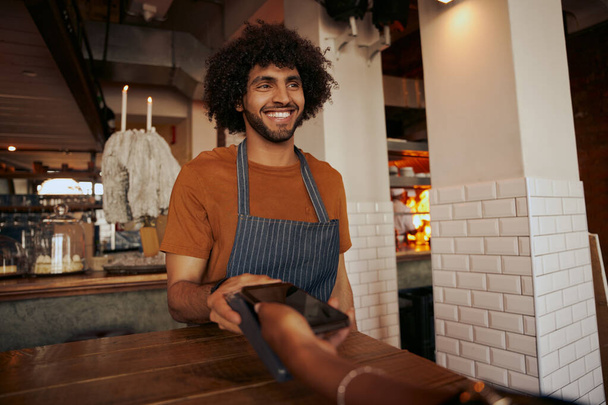 Young man barista wearing casual clothes with apron holding cashless terminal to pay for order at the counter for customer using smartphone - Foto, Imagem