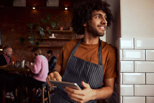 Portrait of a happy waiter standing at restaurant leaning on wall holding digital tablet - Foto, Bild